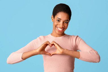 Pozitif Afro-Amerikalı kadın kalp hareketi yapıyor, elleri göğsüne yakın, aşk işareti yapıyor ve kameraya gülümsüyor, siyah kadın hayranlığını dile getiriyor, mavi stüdyo arka planında duruyor, boşluğu kopyalıyor.