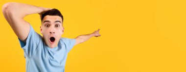 Yakışıklı genç adam şok içinde kafasını tutuyor, ağzı açık boş bir yeri işaret ediyor, büyük indirimler sunuyor, turuncu stüdyo arka planında ürününüzün reklamını yapıyor. Panorama