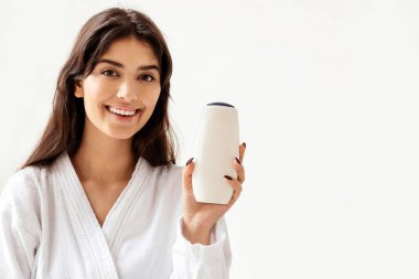 Bornozlu kadın şampuan şişesi reklamı yapıyor. Güzellik ürünleri. Evdeki banyoda, küvette oturan kameraya gülümsüyor. Spa ve Vücut Bakımı Kozmetik Konsepti. Duş jeli Reklamı