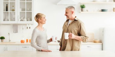 Günaydın, harika nazik ruh hali, taze kahve içkisi ve evde birlikte kahvaltı. Orta yaşlı mutlu bayan ve erkek, modern mutfağın içinde bir bardak sıcak içecekle, günışığında, boşluğu kopyalayın.