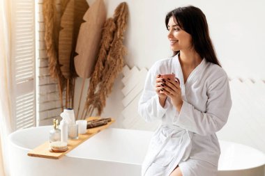 Modern banyodaki banyo küvetinde sabah kahvesi içen mutlu bayan. Beyaz bornoz giyen, spa gününün tadını çıkaran, evde yıkandıktan sonra kahve fincanıyla poz veren bir kadın. İyilik ve Güzellik