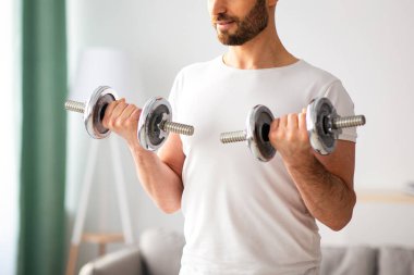 Yakışıklı orta yaşlı adamın evde dambıl egzersizi yapması, kol gücü üzerinde çalışması, pazularına bakması, fotokopi çekmesi. Atletik adam oturma odasının iç tarafına dambılları kaldırıyor.