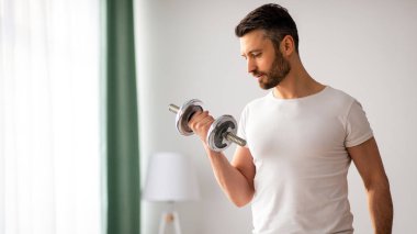 Yakışıklı orta yaşlı adamın evde dambıl egzersizi yapması, kol gücü üzerinde çalışması, pazularına bakması, fotokopi çekmesi. Atletik adam oturma odasının iç tarafına dambılları kaldırıyor.