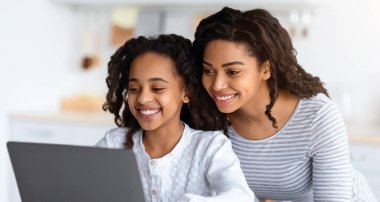 Gülümseyen Afro-Amerikan annesi çocuğuna yardım ediyor, bilgisayar ekranına bakıyor, fotokopi çekiyor. Diz üstü bilgisayar kullanan mutlu siyah kız, koronavirüs salgını, mutfak içi...