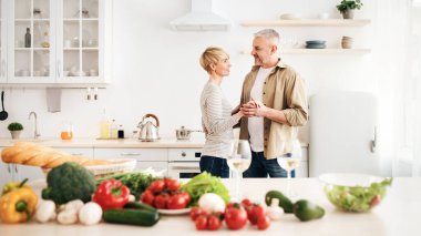 Rahat bir dairede romantik bir akşam yemeği, boş zamanın ve sağlıklı bir yaşam tarzının tadını çıkar. Neşeli son sınıf öğrencisi erkek ve bayan açık mutfak içlerinde açık renkli sebzelerle dans ediyor.