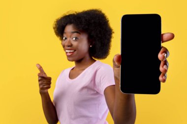 Uygulama ekranı ya da metin için bir model. Sarı stüdyo arka planında boş cep telefonu ekranı gösteren Afrikalı Amerikalı bir kadın. Siyah kadının elinde boş ekranlı akıllı telefon, yakın plan.