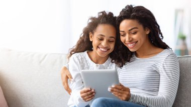 Mutlu Afro-Amerikan anne ve çocuğu evdeki kanepede oturup fotokopi çekerken dijital tablet kullanıyorlar. Siyah anne ve kızı seviyorlar internette oyunlar oynuyorlar ve sarılıyorlar.