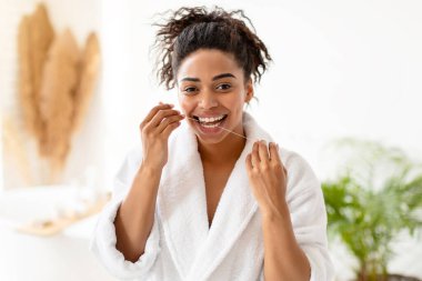 Diş ipi kullanan, diş ipi kullanan, kameraya gülümseyen ve günlük diş bakımının keyfini çıkaran, modern banyoda dikilen, bornoz giyen Afrikalı Amerikalı bir kadın. Diş Bakımı Konsepti