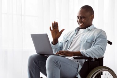 Çevrimiçi sohbet, video görüşmesi ve ev hapsi sırasında görüşme. Keyifli, olgun, Afro-Amerikan tekerlekli sandalyedeki engelli adam oturma odasında ya da klinikte dizüstü bilgisayar ve el sallıyor.