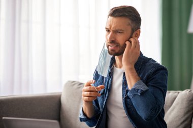 Sakallı, orta yaşlı, koruyucu yüz maskesi takan, kaşındıran sakalını kaşıyan, Coronavirus salgını sırasında laptopta çalışan serbest çalışan, kopyalama uzayı olan bir adam. Yüz maskesi cildi sorunları kavramı