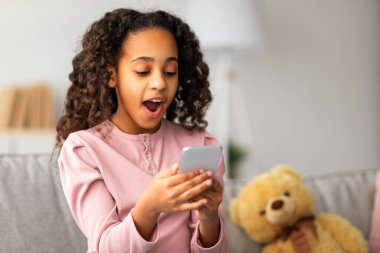 Şok içeriği. Şaşırmış Afro-Amerikan kız akıllı telefona bakıp, iyi haberler alıyor. Kanepede oturan neşeli genç, evde dinleniyor, boş yer var.
