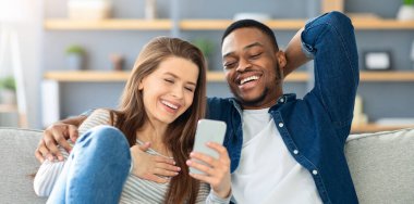 Neşeli Irklar Arası Aşıklar Evde Smartphone 'da komik videolar izliyor, Mutlu Çok Kültürlü Çiftler Oturma odasında dinleniyor, gülüyor ve birlikte vakit geçiriyorlar,