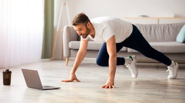 Pozitif atletik sakallı orta yaşlı adam evde egzersiz yapıyor, dizüstü bilgisayar kullanıyor, internette spor videoları izliyor, fitness dersi alıyor, yer kopyalıyor. Sağlıklı yaşam tarzı, kendini soyutlama sporu.