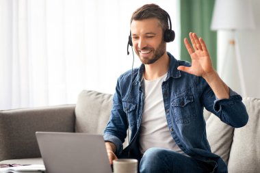 Gülümseyen sakallı iş adamı evden video konferansı yapıyor, kulaklık ve laptop kullanıyor, konuşuyor ve el kol hareketi yapıyor. Keyifli orta yaşlı bir adam oturma odasında koltukta oturuyor, görüntülü konuşma yapıyor, boşluğu kopyalıyor.