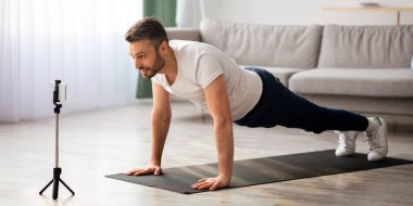 Fitness minderinde motivasyonlu orta yaşlı adam eğitimi, akıllı telefonla yayın, evde otururken spor eğitmeniyle çevrimiçi görüşme, yer kopyalama. Koç cep telefonuyla video çekiyor.