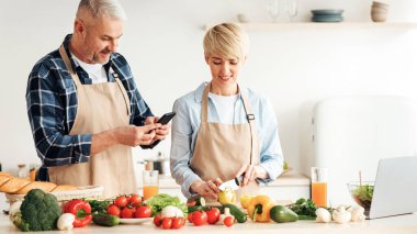 Birlikte lezzetli yemekler, sağlıklı yemekler ve vejetaryen yemekleri pişirin. Önlüklü mutlu bir son sınıf kadını yemek hazırlar, adam akıllı telefonuyla fotoğraf çeker mutfağın içinde parlak sebzeler ve meyve suyu ile.