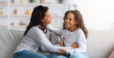 Neşeli genç siyah anne ve kız evde, kenar manzarada, kopyalama alanında birlikte eğleniyorlar. Mutlu Afro-Amerikan annesi oturma odasındaki kanepede birlikte otururken gülen kızını gıdıklıyor.