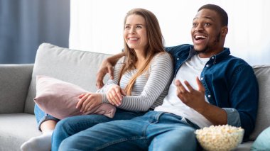 Hafta sonu tatili. Neşeli, ırklar arası bir çift TV seyrediyor ve evdeki koltukta dinleniyor, mutlu, genç, çok kültürlü aşıklar sıcak oturma odasında dinleniyor ve patlamış mısır yiyor, yakın plan çekimler yapıyor.