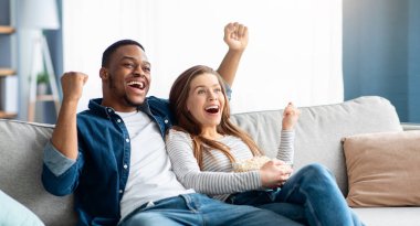 Ev Eğlencesi. Mutlu ırklar arası çiftler televizyon izleyip, evde tezahürat yapıyor, çok ırklı aşıklar patlamış mısır yiyor ve oturma odasında koltukta dinlenirken heyecanla bağırıyor.