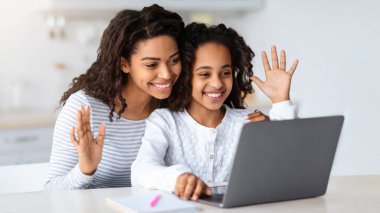 Şirin siyah anne ve çocuğun öğretmeniyle görüntülü konuşması, dizüstü bilgisayara el sallaması ve gülümsemesi, beyaz mutfak içi, fotokopi alanı. Neşeli Afrikalı Amerikalı kız ve annesi babamla görüntülü konuşma yapıyorlar.