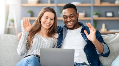 Evdeki dizüstü bilgisayarında video görüşmesi yapan, oturma odasında otururken web kamerasına el sallayan, siyahi erkek ve beyaz kadının internet iletişiminin keyfini çıkardığı mutlu ırklar arası çift.