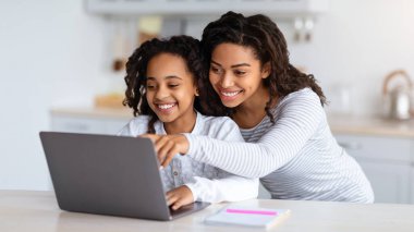 Gülümseyen Afrikalı Amerikalı anne çocuğunu kucaklıyor, bilgisayar ekranına bakıyor, fotokopi çekiyor. Diz üstü bilgisayar kullanan mutlu siyah kız, koronavirüs salgını, mutfak içi...