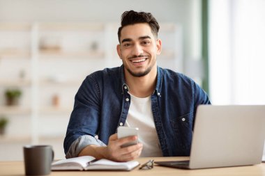 Gülümseyen Arap adam evde çalışma masasında otururken akıllı telefon kullanıyor, dizüstü bilgisayar üzerinde çalışıyor, kopyalama alanı kullanıyor. Elinde cep telefonu olan neşeli, yakışıklı, orta doğulu bir adam.