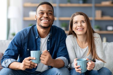 Hafta sonu tatili. Neşeli, genç, çok kültürlü bir çift oturma odasında dinleniyor, mutlu ırklar arası aşıklar televizyon seyrediyor ve evde kahve içiyor, ev içi dinlenmenin keyfini çıkarıyor, yakın çekim yapıyor.
