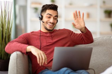 Evde çalışan, dizüstü bilgisayarlı koltukta oturan, kulaklık kullanan pozitif Arap bir adam. Ortadoğulu bir adam müşterileri ya da iş arkadaşlarıyla görüntülü görüşme yapıyor.