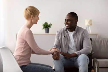 Her şey yoluna girecek. Evde ya da klinikte aile desteği ve sağlık hizmetleri. Neşeli, dost canlısı, sakin, Kafkasyalı kadın, oturma odasında sakat kalmış yetişkin bir Afrikalı Amerikalının elini tutuyor.