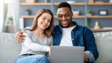 İnternette dizüstü bilgisayarı olan mutlu, ırklar arası bir çift, mutlu, çok kültürlü eşler bilgisayarla koltukta dinleniyor, internetten alışveriş yapıyor ya da tatil ayarlıyor,