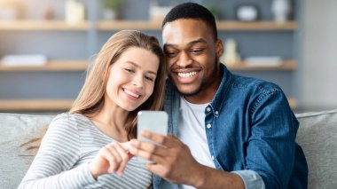 Neşeli çift akıllı telefonlarıyla evde vakit geçirir, video izler veya internetten cep telefonuyla alışveriş yapar, ırklar arası eşler oturma odasında kanepede dinlenir, yakın çekim yapar.