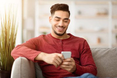 Mutlu Arap adam yeni mobil uygulamanın tadını çıkarıyor, elinde akıllı telefon ve gülümsüyor. Neşeli orta doğulu adam evdeki koltukta uzanırken cep telefonu kullanıyor arkadaşlarıyla sohbet ediyor, mekanı kopyalıyor.