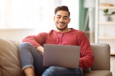 Gülümseyen yakışıklı Arap adam koltukta oturuyor ve evinde dinlenirken modern dizüstü bilgisayar kullanıyor. Neşeli orta doğulu genç adam defter kullanıyor, kız arkadaşıyla sohbet ediyor ya da web sörfü yapıyor.