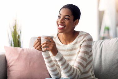 Hafta sonu tatili. Neşeli, genç, siyah ev hanımı, oturma odasında rahat bir koltukta dinlenirken kahve içiyor, elinde sıcak içecekle gülümseyen ve uzaklara bakan Afrikalı Amerikalı bir kadın.