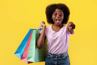 Hey, sen! Duygusal siyahi kadın kamerayı işaret ediyor ve sarı arka planda duran renkli alışveriş poşetleri tutuyor, stüdyo çekimi, boş alan. Alışveriş ve satış zamanı.