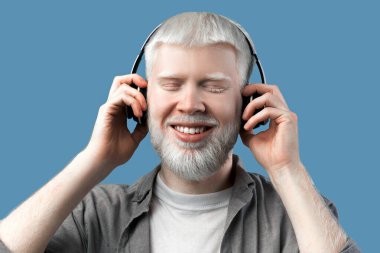 Kablosuz kulaklıkla müzik dinleyen ve turkuaz arka planda onlara dokunan mutlu albino adam. Huzurlu adam gözlerini kapatıyor ve gülümsüyor, favori melodilerin tadını çıkarıyor.