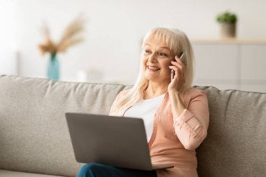 Gülümseyen olgun bir kadının portresi cep telefonuyla konuşuyor, bilgisayarda çalışıyor, oturma odasında kanepede oturuyor ve başka tarafa bakıyor. Telekomünikasyon ve telesekreter. Dizüstü bilgisayar kullanan kendine güvenen yetişkin
