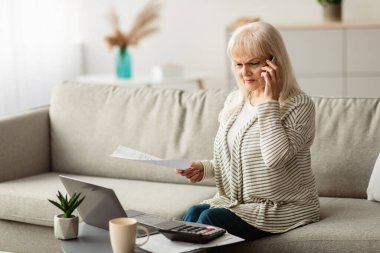 Evrak işi kavramı. Evde dizüstü bilgisayarla çalışan, cep telefonuyla konuşan, evrakları tutan, proje ya da belgenin detaylarını tartışan, evdeki ofiste koltukta oturan ilgili olgun bir kadının portresi.