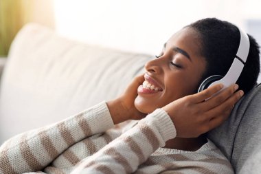Rahatlama zamanı. Genç, kablosuz kulaklıklı, müzik ya da Audiobook dinleyen, koltukta uzanan, gülümseyen, Afro-Amerikan kadın boş zamanların tadını çıkaran, yakın çekim yapan,