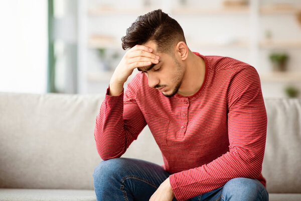 Closeup portrait of pensive bearded arab guy having difficulties, sitting on couch at home, touching his head and looking down, panorama with copy space. Financial hangover while pandemic concept