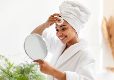 Pamuk pedli ve aynalı siyahi kadın yüz bakımı yapıyor. Evde tuvalette makyaj yapıyor. Kapalı alanda yüz derisini önemseyen kadın. Güzellik Bakımı Konsepti. Seçici Odaklanma