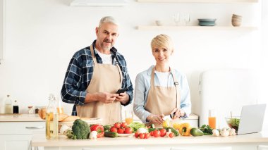 Yemek blogcusu, yemek dersi, covid-19 karantinasından kendini soyutlama. Yetişkin, neşeli eş salata yapar. Mutfakta akıllı telefonu olan bir adam. Masada taze, parlak sebzeleri olan bir adam.