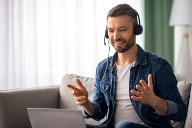 Neşeli, yakışıklı sakallı adam evden video konferansı yapıyor. Oturma odasında otururken kulaklık ve laptop kullanıyor. İş ortaklarıyla konuşuyor, gülümsüyor ve el kol hareketi yapıyor.