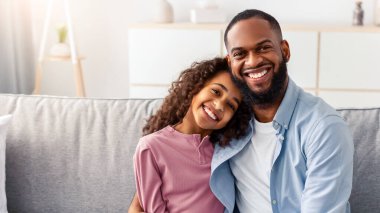 Bağ ve Birliktelik Konsepti. Afrikalı Amerikalı baba ve kızın portresi kucaklaşıp gülümsüyor, birlikte eğleniyorlar. Mutlu adam bir kıza sarılıyor, oturma odasında kanepede oturuyor.