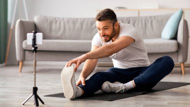 Keyifli, orta yaşlı sporcu bir adam fitness minderinde geriniyor, akıllı telefonla yayın yapıyor, spor eğitmeniyle çevrimiçi görüşme yapıyor evde otururken, yer kopyalıyor. Koç cep telefonuyla video çekiyor.