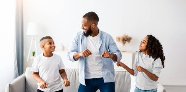 Parenthod ve Mutlu Aile Kavramı. Heyecanlı kızı ve oğluyla müzik eşliğinde dans eden pozitif siyah baba. Eğlenceli çocuklar babalarıyla eğleniyor, boş zaman ve hafta sonları oturma odasında eğleniyorlar.