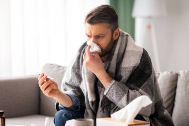 Orta yaşlı sakallı bir adam battaniyeye sarılmış, kendini hasta hissediyor, ateşi termometreyle kontrol ediyor, hapşırıyor, ev içi, fotokopi odası. Soğuk algınlığı, grip veya koronavirüs enfeksiyonu konsepti