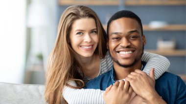 Mutlu Çok Kültürlü Çift Portresi. Siyahi erkek ve beyaz kadının evde kucaklaşması, kameraya gülümseyen ve kucaklaşan ırklar arası aşıklar, evdeki kanepede dinlenirken,