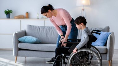 Genç anne tekerlekli sandalyedeki özürlü oğluna bacağını evde oynatması için yardım ediyor. Engelli çocuğuyla ilgilenen bin yıllık bir ebeveyn.